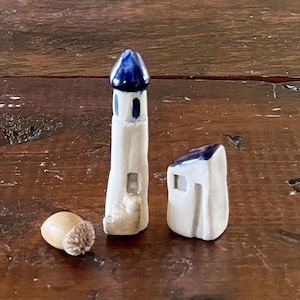 Pode incluir: Uma miniatura de farol de cerâmica com um topo azul e um pequeno edifício de cerâmica com um telhado azul. Uma bolota está ao lado das estruturas de cerâmica. Os itens estão em uma superfície de madeira.