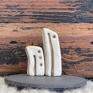 May include: Two small white ceramic sculptures of buildings with brown accents. The sculptures are sitting on a black slate base.