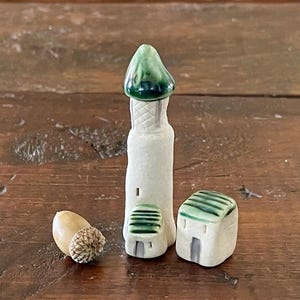 May include: Three small ceramic houses with green roofs. The tallest house has a pointed roof and the other two have flat roofs. The houses are white with green accents. There is a small acorn in the foreground.