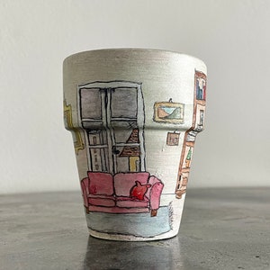 May include: A white ceramic planter with a hand-painted scene of a living room interior. The scene includes a couch, a window, a bookshelf, and artwork on the walls.