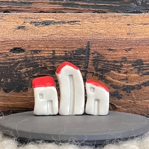 Puede incluir: Tres pequeñas casas de cerámica blanca con techos rojos, dispuestas en fila sobre una base de pizarra negra. Las casas son de diseño sencillo, con una sola ventana y una pequeña puerta.
