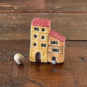May include: A miniature ceramic building with a red roof and yellow walls. The building has multiple windows and a small door. The building is sitting on a wooden surface.