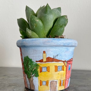 Peut inclure: Un pot en terre cuite peint à la main avec un glaçage bleu et blanc représente une scène fantaisiste d'une maison jaune avec des volets orange et une maison rouge avec une porte blanche. Un arbre vert se trouve devant la maison jaune. Le pot est rempli d'une plante succulente verte.
