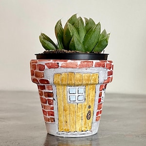 Può includere: Vaso di fiori in terracotta dipinto a mano con un design stravagante. Il vaso presenta una facciata in mattoni con una porta gialla e una piccola finestra. All'interno c'è una pianta succulenta verde.