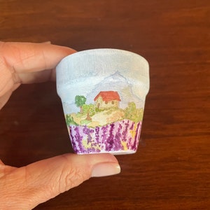 May include: A small white ceramic flower pot with a painted scene of a house with a red roof, a green tree, and purple flowers. The pot is decorated with a watercolor style.
