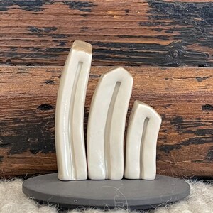 May include: Three white ceramic sculptures in the shape of tall, thin, rounded rectangles, arranged in descending size order on a black circular base. The sculptures have a smooth, glossy finish.
