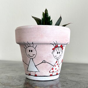 May include: A pink terracotta planter with a hand-painted design of two stick figures holding hands. The figures are a girl with a red bow and a boy with a white shirt. The planter has a small succulent plant growing in it.