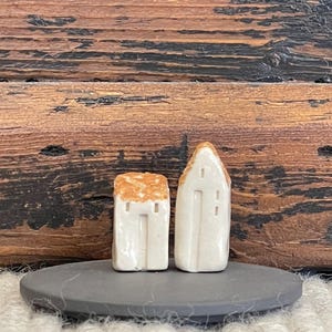 Op de afbeelding: Twee kleine witte keramische huisjes met bruine daken, die op een zwarte leisteenbasis staan. De huisjes hebben eenvoudige details, waaronder ramen en deuren.