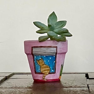 May include: A pink painted terracotta planter with a hand-painted design of a cat looking out a window. The planter has a succulent plant growing in it.