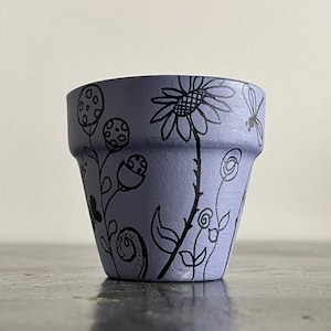 Puede incluir: Una pequeña maceta de color lavanda con diseños florales negros dibujados a mano. La maceta presenta un girasol, otras flores y una libélula. La maceta está sobre una superficie oscura.