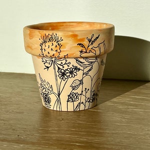 Peut inclure: Un petit pot de fleurs rond en terre cuite avec un extérieur pêche clair et un bord pêche plus foncé. Le pot est décoré de dessins au trait noirs de diverses fleurs et feuillages. Le pot est posé sur une surface en bois.