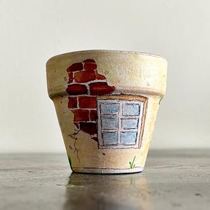 Può includere: Un piccolo vaso di fiori in terracotta dipinto a mano. Il design presenta un muro di mattoni e una finestra su uno sfondo color crema. Il vaso ha un aspetto invecchiato, con crepe e vernice scrostata, ed è adatto a piccole piante.