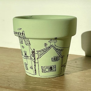 May include: A light green ceramic flower pot with a hand-drawn black illustration of houses, chimneys, and power lines. The pot has a smooth, matte finish and is suitable for indoor or outdoor use.