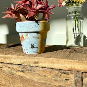 May include: A terracotta pot with a hand-painted design of a building with laundry hanging on a clothesline. A black cat sits on a ledge. The pot is filled with a succulent plant with red leaves.