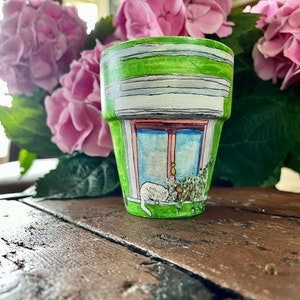 May include: A green painted flower pot with a white window and a white cat sleeping in the window. The window has a pink frame and a blue sky is visible through the window.