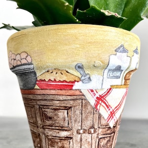 Può includere: Un vaso di fiori in ceramica giallo con un design dipinto a mano di un bancone da cucina con una torta, una ciotola di uova, un contenitore di farina e un mattarello. Il vaso ha una tovaglia a quadri rossi e bianchi.