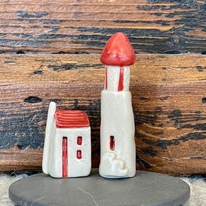 May include: Two ceramic figurines: a small house and a lighthouse. The house has a red roof and a white body with a red door and windows. The lighthouse is white with red accents and a red top. Both are on a gray, round base.