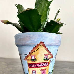 Könnte beinhalten: Ein kleiner Terrakotta-Topf, bemalt mit einem gelben Haus mit einer rosafarbenen Tür und dem Schriftzug "Free Love" auf einem Schild. Der Topf ist mit einer grünen Kaktuspflanze mit rosa Knospen gefüllt.