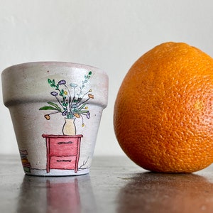 May include: A small terracotta flower pot with a hand-painted design of a vase of flowers and a small red dresser. The pot is sitting on a gray surface next to a large orange.