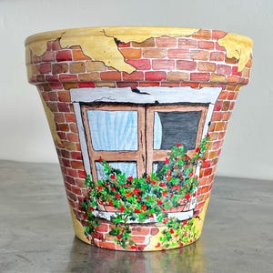 May include: Hand-painted terracotta flower pot featuring a brick wall design with a window and flowers. The pot has a yellow rim and a weathered appearance, adding a rustic charm. The window has blue curtains and a dark interior.