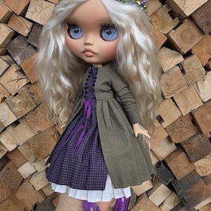 Può includere: Una bambola con lunghi capelli ondulati biondo platino e grandi occhi azzurri. Indossa una corona di perline nei toni del viola e del verde, un cappotto verde oliva scuro sopra un vestito viola a motivi con sottogonna bianca e scarpe viola.