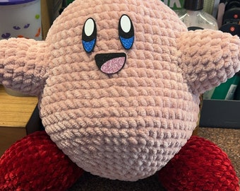May include: A pink and red crocheted plush toy with a smiling face and blue eyes. The toy is sitting on a brown surface.