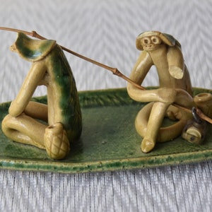 May include: Two ceramic figurines of monkeys sitting in a green leaf-shaped boat, holding fishing rods. The monkeys are wearing hats and have detailed facial features.