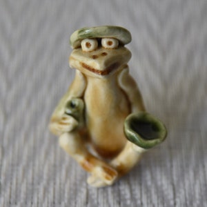 May include: A small ceramic figurine of a frog wearing a green hat and holding a cup. The frog is painted in shades of brown, green, and white.