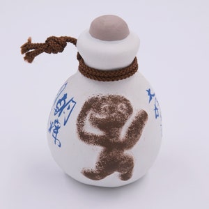 May include: A white ceramic bottle with a brown rope tied around the neck. The bottle has a brown figure painted on the front and blue Chinese characters on the side.