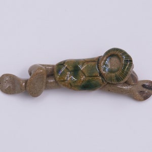 May include: A ceramic turtle figurine in shades of brown and green. The turtle has a textured shell with a hexagonal pattern and a coiled head. The figurine is lying down, with its legs extended.
