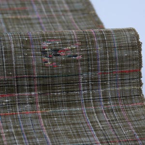 Pode incluir: Close-up de um tecido texturizado com um padrão xadrez. O tecido é principalmente verde-oliva com uma grade de linhas brancas, vermelhas, azuis e pretas. A borda do tecido está desfiada.