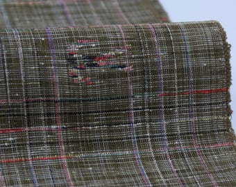 May include: Close-up of a textured fabric with a plaid pattern. The fabric is primarily olive green with a grid of white, red, blue, and black lines. The edge of the fabric is frayed.