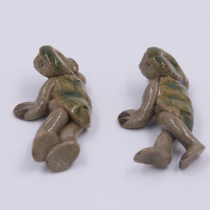 May include: Two ceramic turtle figurines in a relaxed pose. The turtles have a light brown body with green and brown shell details. The figurines are on a white background.