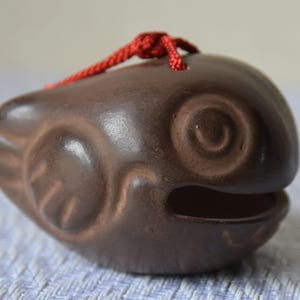Peut inclure: Un hochet en céramique marron en forme de poisson avec un cordon rouge. Le hochet présente des détails sculptés pour l'œil, les nageoires et la bouche. Le poisson est de couleur marron foncé.