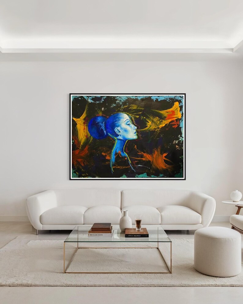 Pode incluir: Uma grande pintura quadrada em uma moldura preta retrata o perfil de uma mulher em tons de azul contra um fundo escuro e abstrato com pinceladas laranja e amarelas. A obra de arte est&aacute; pendurada acima de um sof&aacute; branco em uma sala de estar moderna.