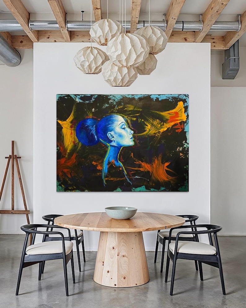 Pode incluir: Pintura abstrata do perfil de uma mulher em tons de azul, amarelo e laranja. A obra de arte est&aacute; pendurada acima de uma mesa de jantar redonda de madeira com cadeiras pretas. V&aacute;rias lumin&aacute;rias pendentes brancas em forma de globo iluminam a sala.
