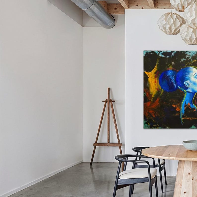 Pode incluir: Uma sala de jantar moderna com uma mesa redonda de madeira e duas cadeiras pretas com almofadas creme. Uma grande pintura com tons de azul e amarelo est&aacute; pendurada na parede. Um cavalete de madeira tamb&eacute;m est&aacute; presente.