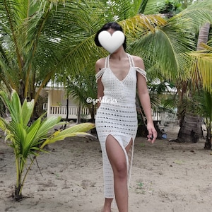 Op de afbeelding: Een witte maxi-jurk van gehaakt kant met een diep decollet&eacute; en splitten aan de zijkanten. De jurk wordt gedragen door een persoon die op een zandstrand staat met palmbomen op de achtergrond.