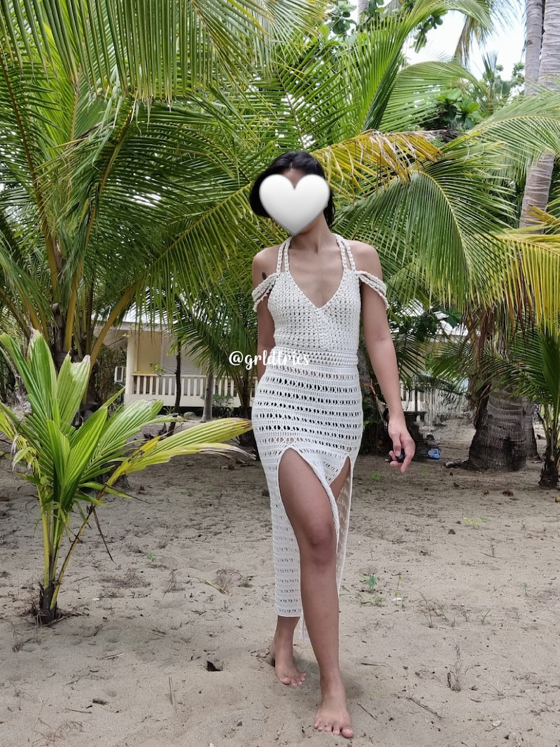 Op de afbeelding: Een witte maxi-jurk van gehaakt kant met een diep decollet&eacute; en splitten aan de zijkanten. De jurk wordt gedragen door een persoon die op een zandstrand staat met palmbomen op de achtergrond.