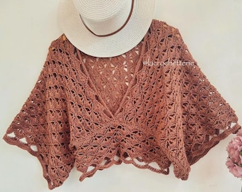 May include: A rust-colored crochet top with a V-neckline and wide sleeves, featuring an intricate openwork pattern and a scalloped hem. A cream-colored straw hat with a brown band rests on top. The text @lacrochetterie is visible on the fabric.
