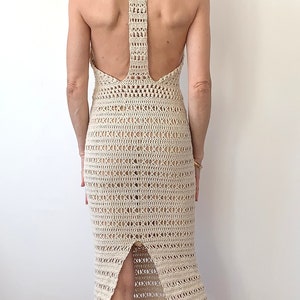 Op de afbeelding: Een witte gehaakte jurk met een halternek en een uitsnijding in de rug. De jurk is aangesloten en heeft een midi-lengte. Het model draagt de jurk met een paar nude hakken.