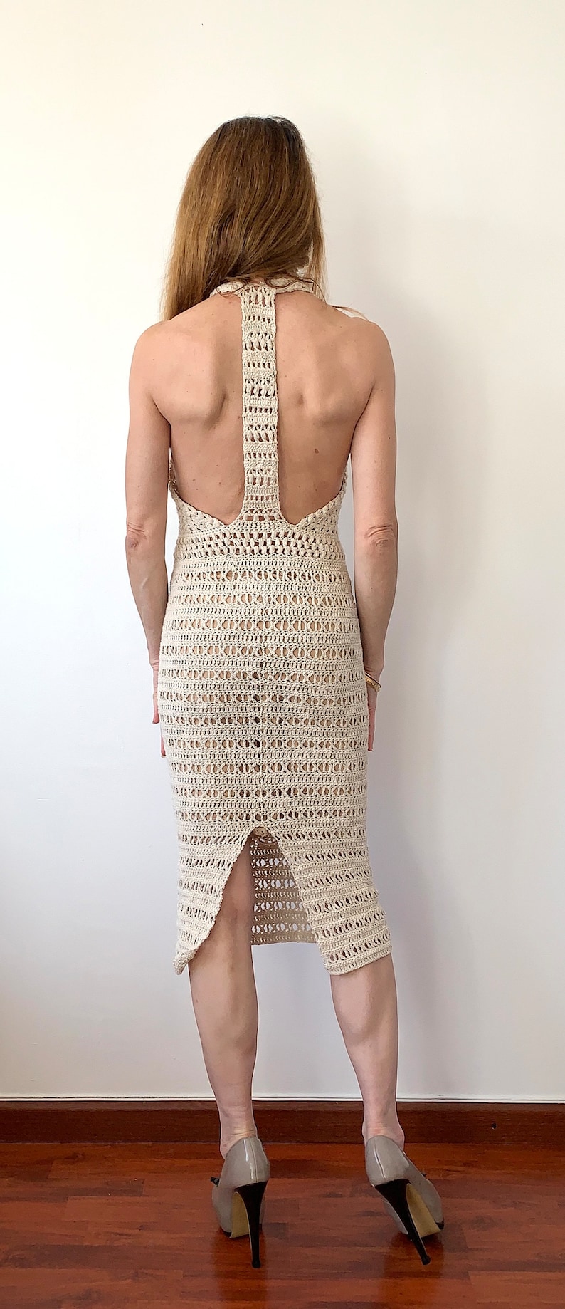 Op de afbeelding: Een witte gehaakte jurk met een halternek en een uitsnijding in de rug. De jurk is aangesloten en heeft een midi-lengte. Het model draagt de jurk met een paar nude hakken.