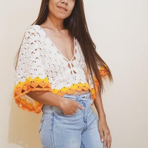 Op de afbeelding: Een wit gehaakt topje met een gele en oranje rand. Het topje heeft een diepe V-hals en korte mouwen. Het model draagt een blauwe jeans.