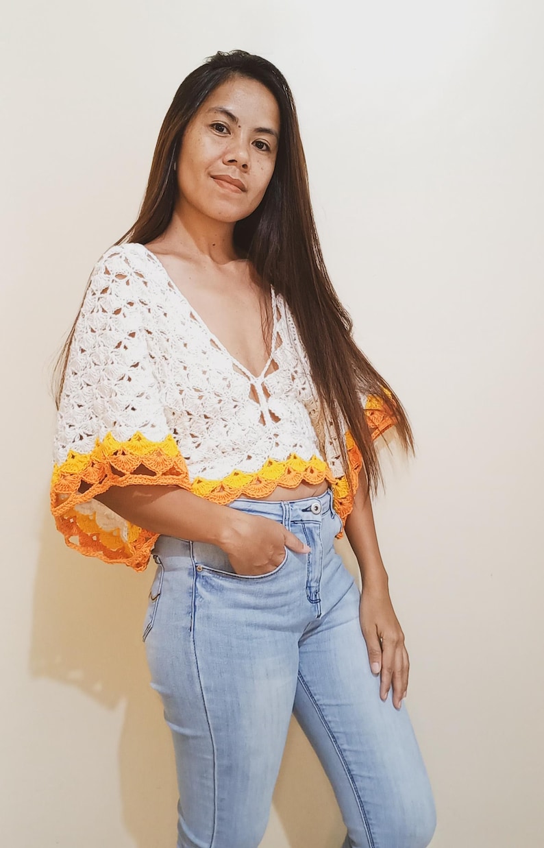 Op de afbeelding: Een wit gehaakt topje met een gele en oranje rand. Het topje heeft een diepe V-hals en korte mouwen. Het model draagt een blauwe jeans.