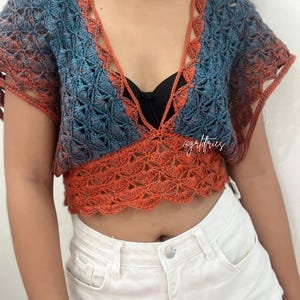 Op de afbeelding: Een gehaakt crop top met een diepe V-hals en korte mouwen. De top is gemaakt in een blauw en oranje ombre patroon. De top wordt gedragen over een zwarte bh.