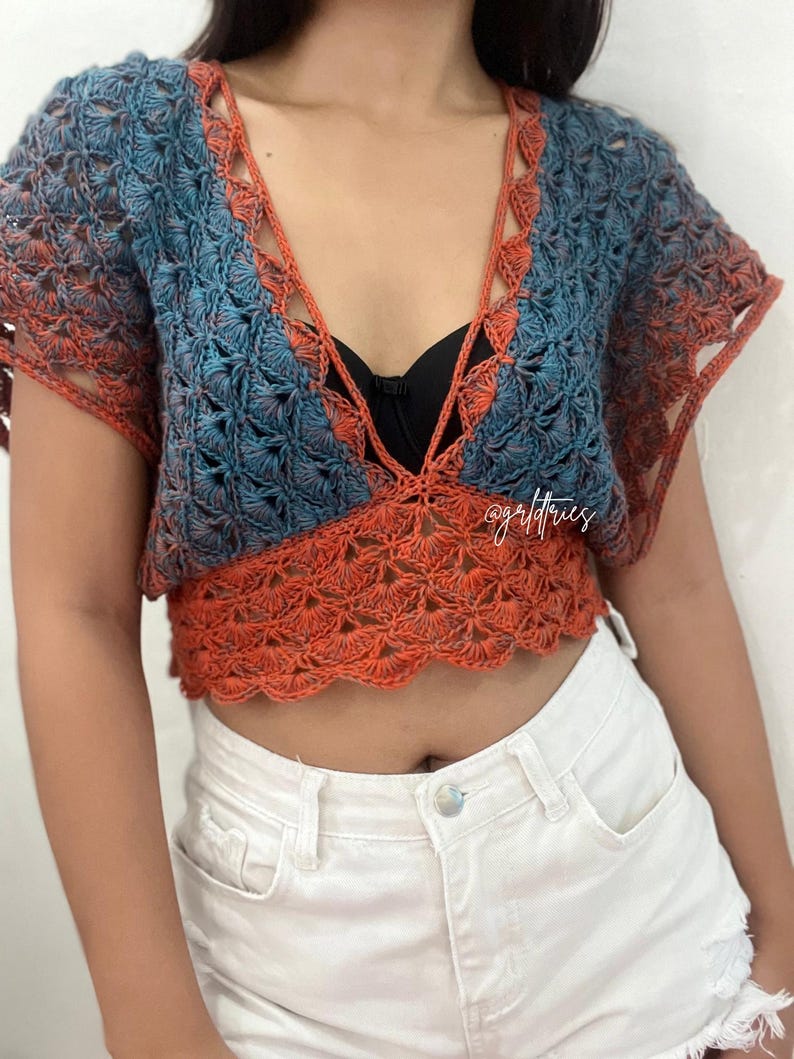 Op de afbeelding: Een gehaakt crop top met een diepe V-hals en korte mouwen. De top is gemaakt in een blauw en oranje ombre patroon. De top wordt gedragen over een zwarte bh.