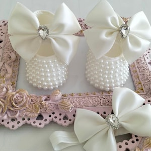May include: A pair of white baby shoes with pearl embellishments and a white satin bow with a rhinestone center. A matching white satin headband with a rhinestone center is also included.