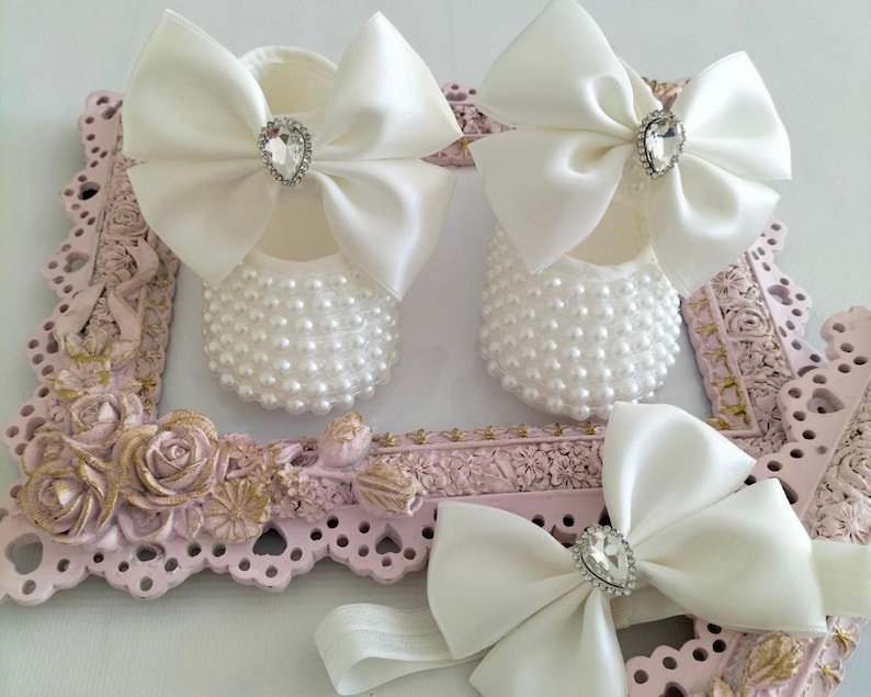 May include: A pair of white baby shoes with pearl embellishments and a white satin bow with a rhinestone center. A matching white satin headband with a rhinestone center is also included.