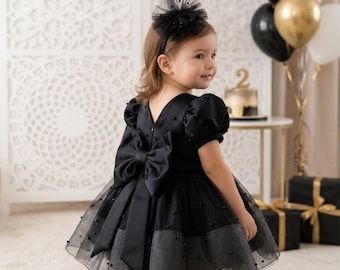 May include: A black dress featuring puff sleeves, a large satin bow at the back, and a tulle skirt overlay adorned with small black pearls. The garment is paired with a matching black headband featuring a tulle flower accent.
