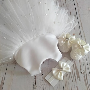May include: White tulle baby dress with pearl embellishments, a matching headband with two satin bows, and a pair of white satin baby shoes with pearl embellishments and satin bows.
